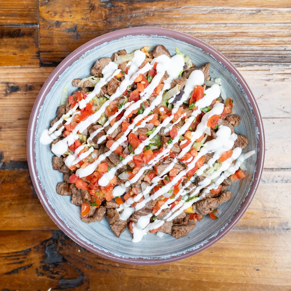 Overhead view of a steak salad with lettuce, diced tomatoes, herbs, and creamy white dressing on a wooden table.