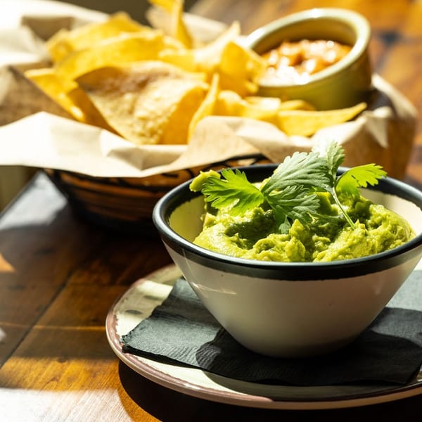 A bowl of guacamole topped with cilantro, with a basket of tortilla chips and salsa in the background.