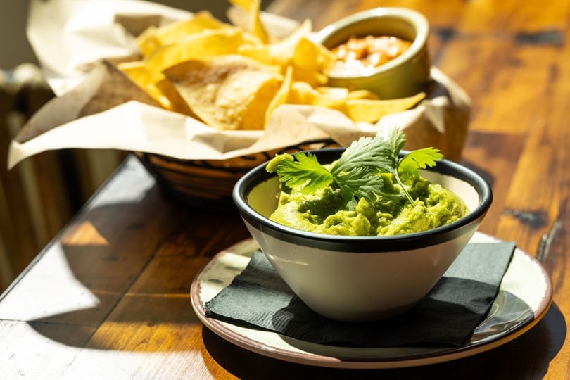 A bowl of guacamole topped with cilantro, with a basket of tortilla chips and salsa in the background.