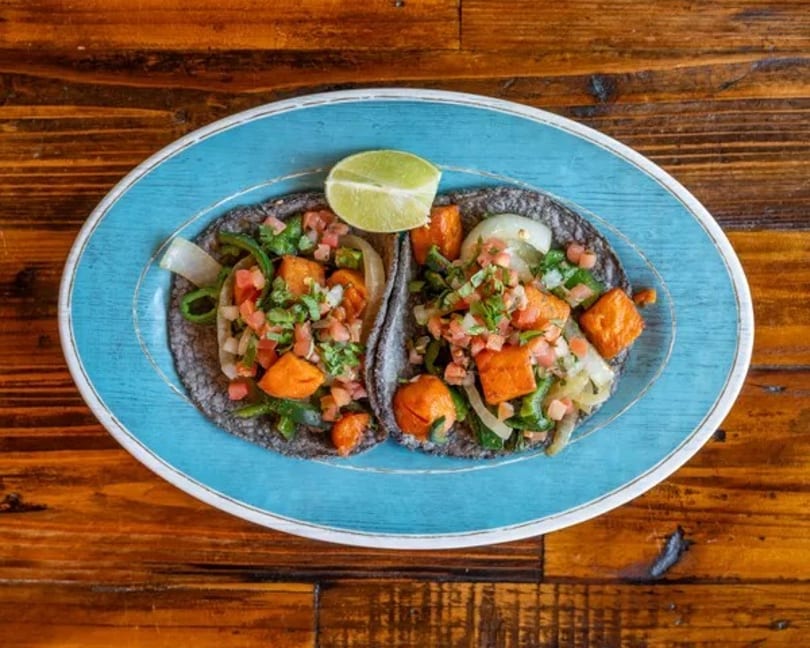 Two vibrant tacos with sweet potato, peppers, and pico de gallo on blue corn tortillas, garnished with lime.