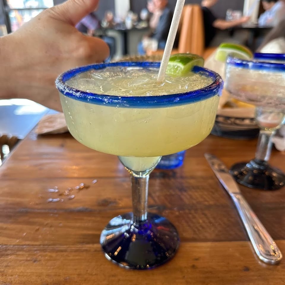Frosted margarita glass with blue rim, lime wedge, and straw on a wooden restaurant table, with diners in the background.
