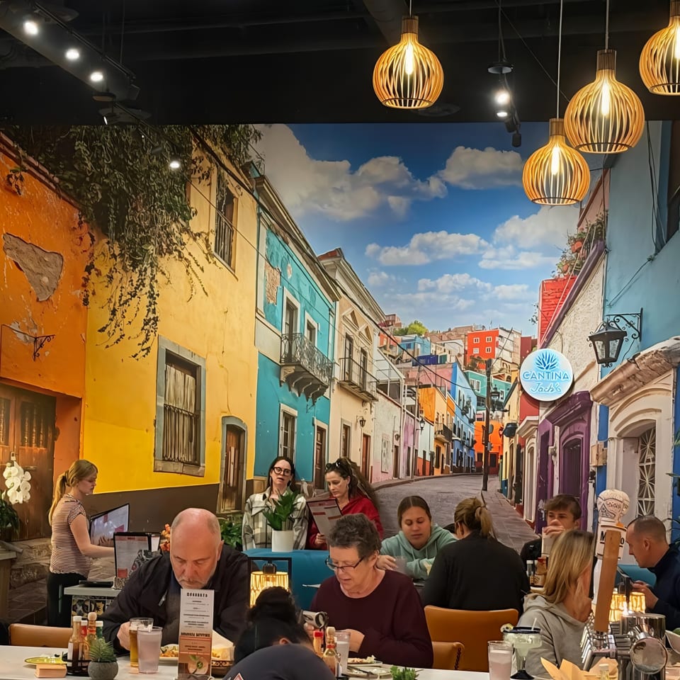 Bustling restaurant dining room with colorful Latin street mural, diners at tables, tropical plants, and woven pendant lights.