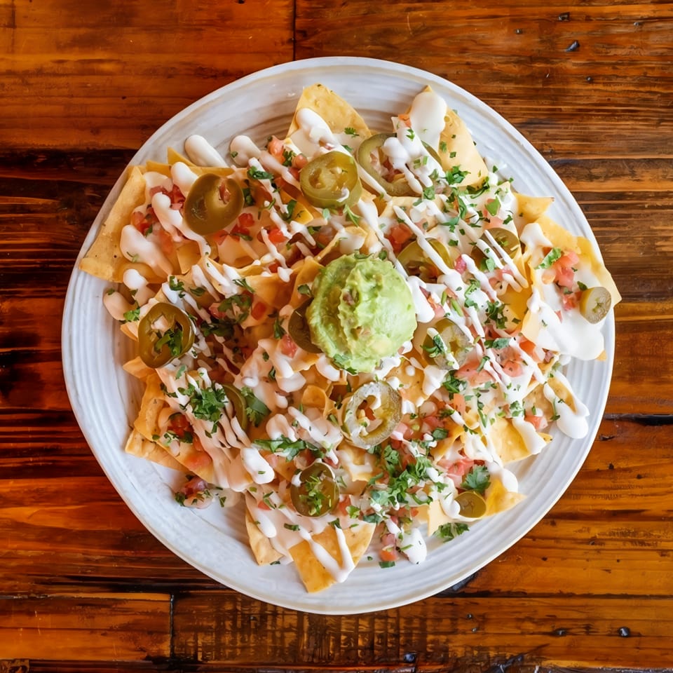 Loaded nachos topped with guacamole, jalapeños, pico de gallo, cilantro, and drizzled cream on a white plate.