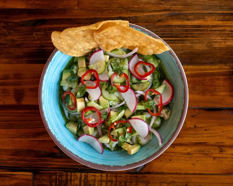 Overhead view of a vibrant ceviche dish with avocado, radishes, and red peppers in a blue bowl.