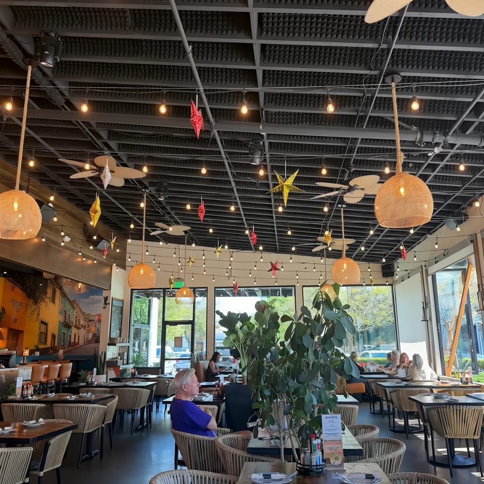 Bright restaurant interior with woven pendant lamps, colorful hanging stars, indoor plants, set tables, and diners by large windows.