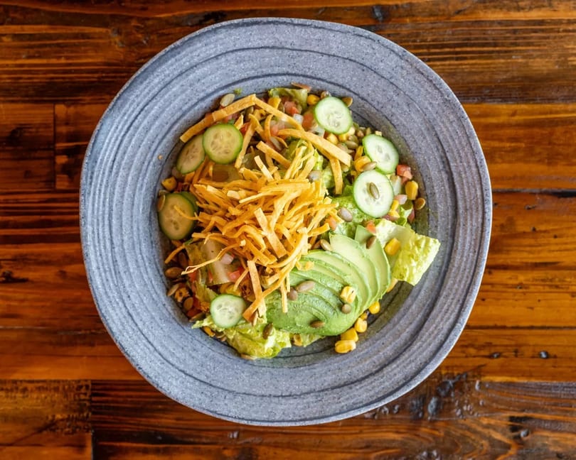Overhead view of a vibrant salad with avocado, corn, cucumbers, and tortilla strips in a gray bowl.