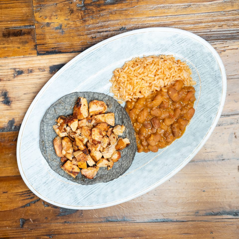 Plated meal featuring grilled chicken on a blue corn tortilla, orange rice, and beans on a rustic wooden table.