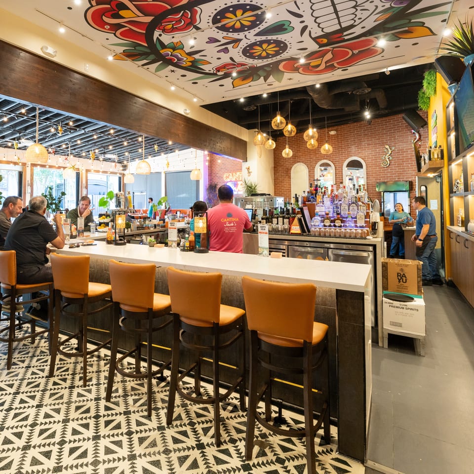 Colorful Cantina restaurant interior with a mural ceiling, patterned floor, bar, counter seating, liquor shelves, and patrons.
