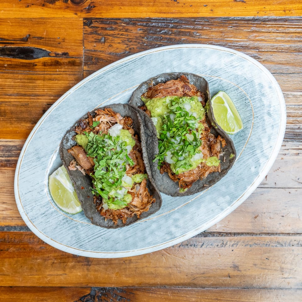 Three shredded-meat tacos on black tortillas with guacamole, cilantro, diced onion, and lime wedges on a blue plate.
