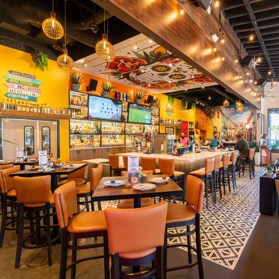 Colorful Latin-inspired restaurant interior with orange seating, a stocked bar, patterned floors, wall murals, and diners.