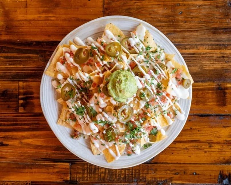 A plate of nachos with guacamole, jalapeños, pico de gallo, and a white sauce drizzle.