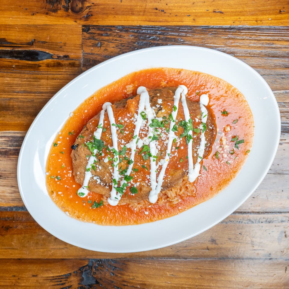 Crispy enchiladas topped with white crema, crumbled cheese, and cilantro in red sauce, served with rice on a white plate.