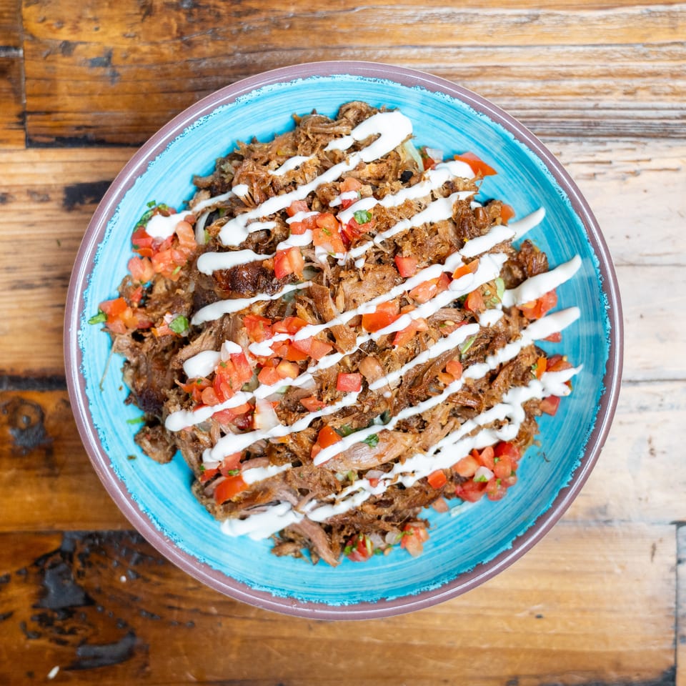 Overhead view of shredded roasted meat topped with diced tomatoes, herbs, and white sauce on a blue plate.