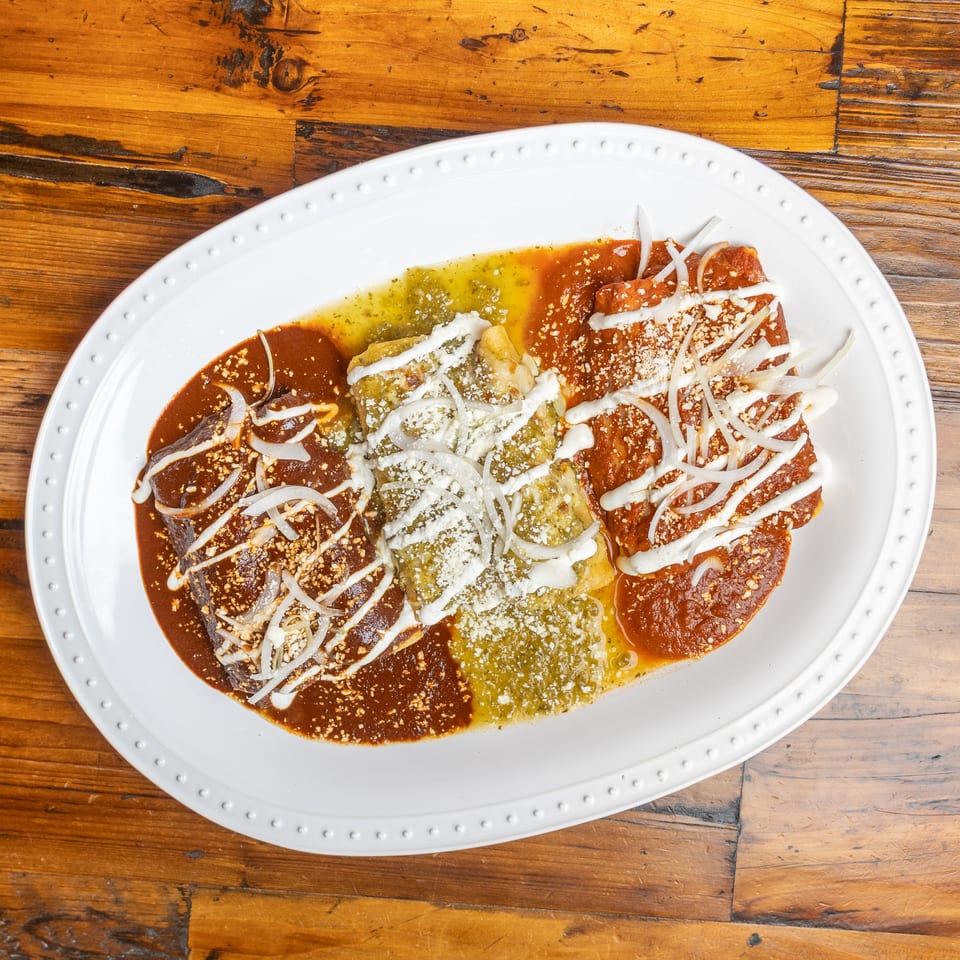 Mexican enchiladas coated in red, green, and brown sauces, topped with onions, cream, and crumbled cheese on a white platter.