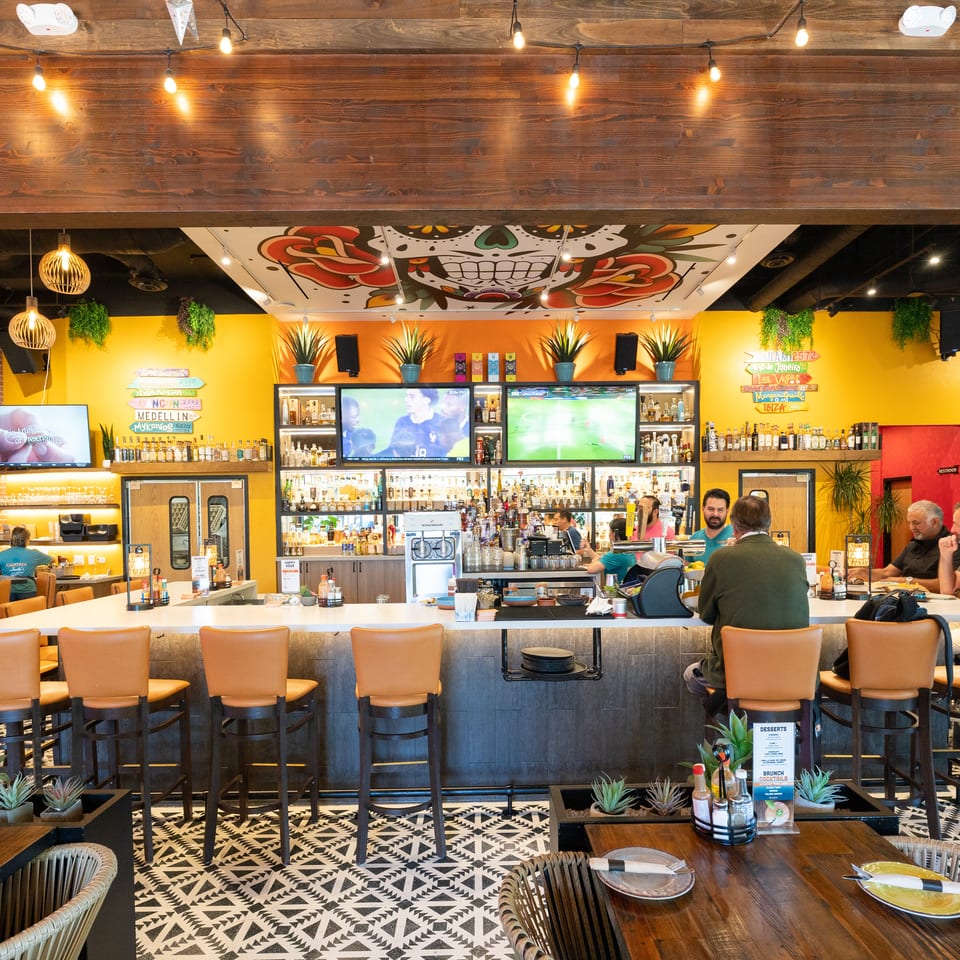 Colorful restaurant bar with liquor shelves, televisions, patterned flooring, skull mural, hanging plants, and seated diners.