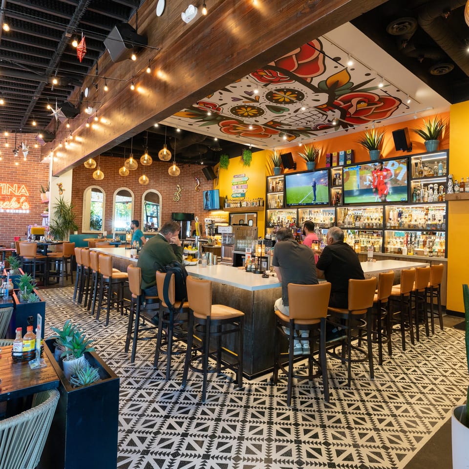 Colorful Cantina Jack’s restaurant interior with a fully stocked bar, televisions, patterned flooring, festive décor, and seated diners.