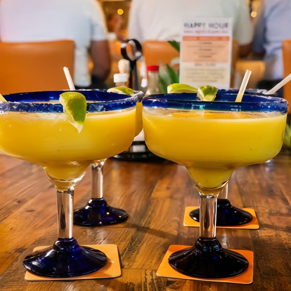 Two yellow margaritas with blue-salt rims and lime wedges sit on orange coasters at a restaurant table.