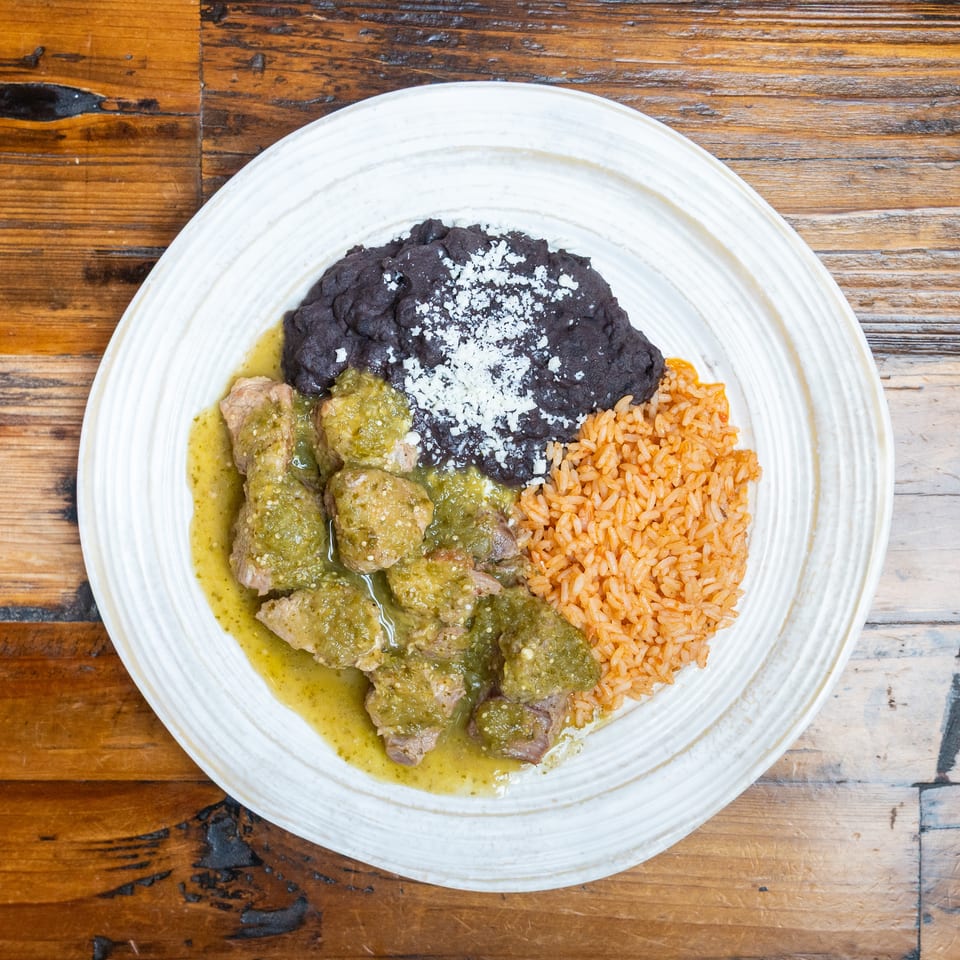 Mexican plate with grilled meat in green salsa, black beans topped with cheese, and orange rice on a wooden table.
