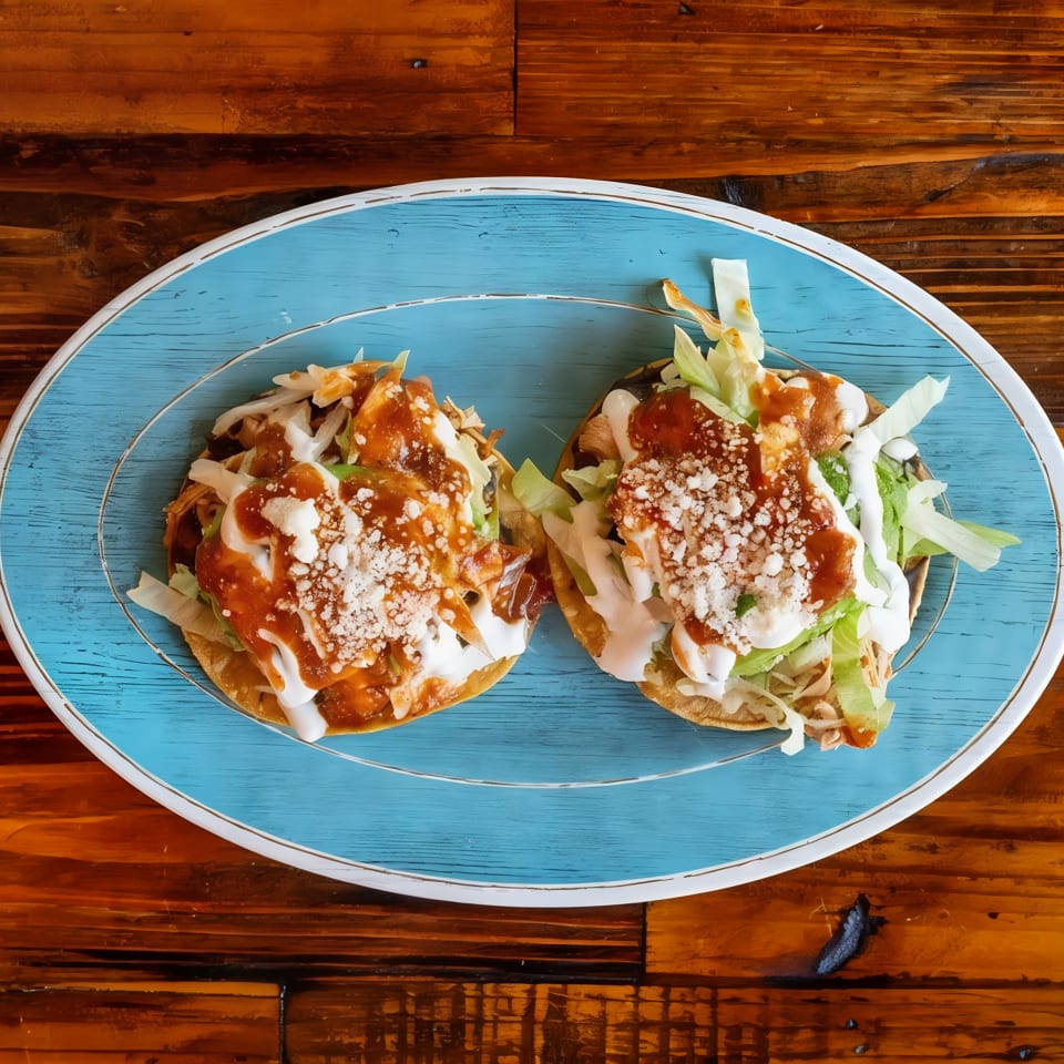 Two loaded tacos topped with shredded lettuce, white sauce, brown salsa, and crumbled cheese on a blue oval plate.
