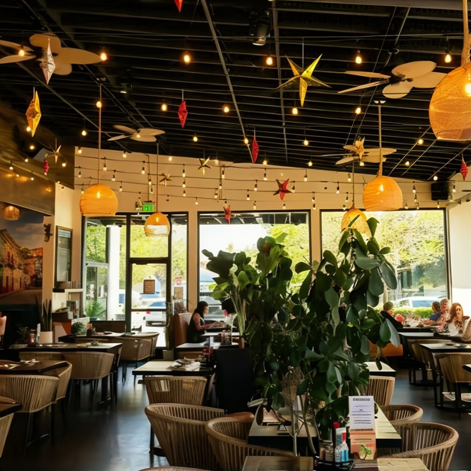 Bright restaurant dining room with leafy plants, woven chairs, pendant lights, hanging ornaments, and patrons seated near large windows.