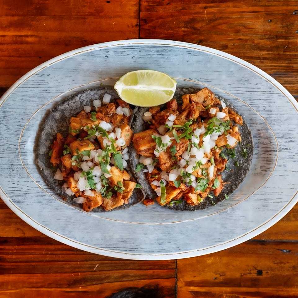 Two black tortillas filled with seasoned meat, diced onions, and cilantro, topped with a lime wedge on a plate.