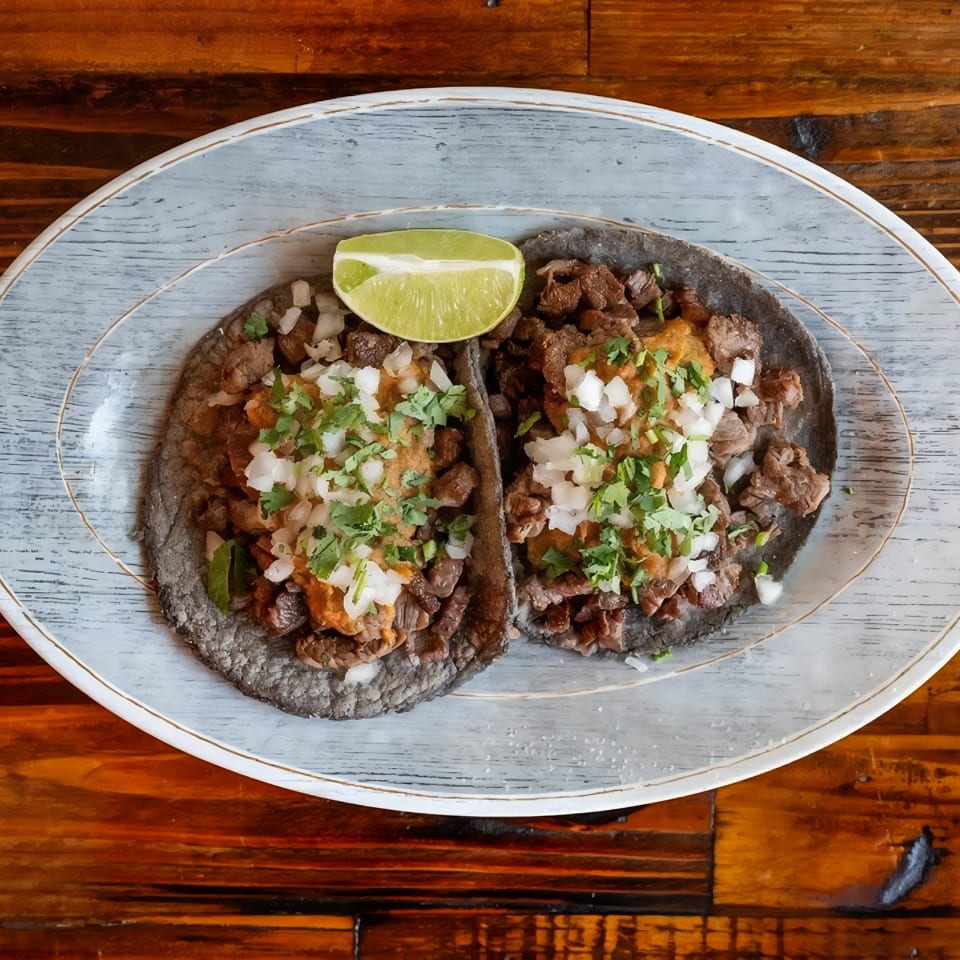 Two tacos filled with seasoned meat, onions, and cilantro, topped with a lime wedge on a ceramic plate.