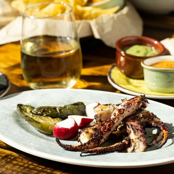 Grilled octopus with roasted peppers and radishes, accompanied by dips and a drink on a wooden table.