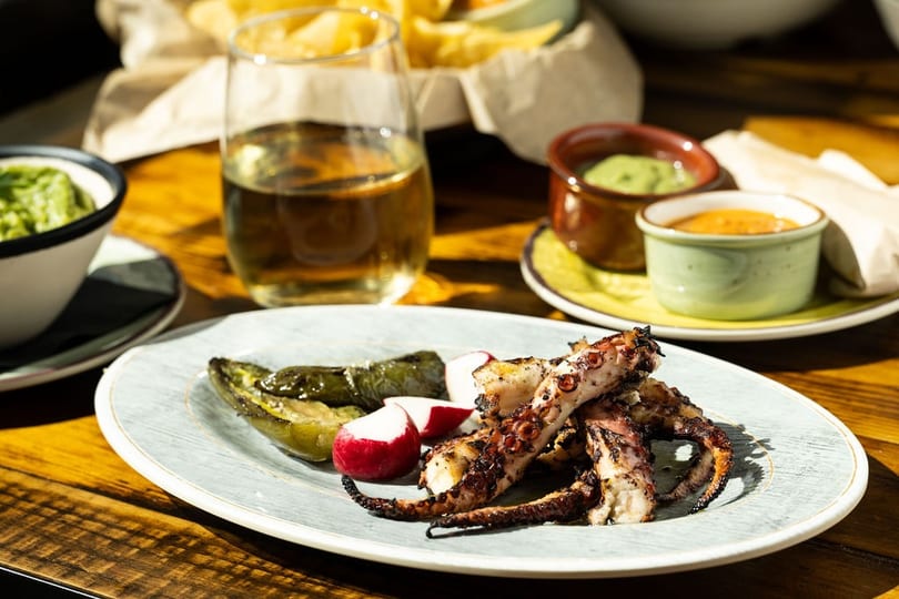 Grilled octopus with roasted peppers and radishes, accompanied by dips and a drink on a wooden table.