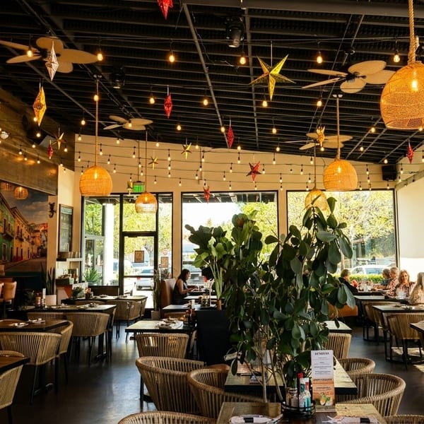 Spacious, vibrant restaurant interior with woven light fixtures, a bar, and patrons dining near large windows.
