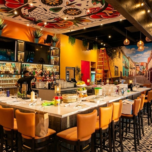 Vibrant Mexican restaurant interior with a long bar, orange stools, and colorful murals on walls and ceiling.