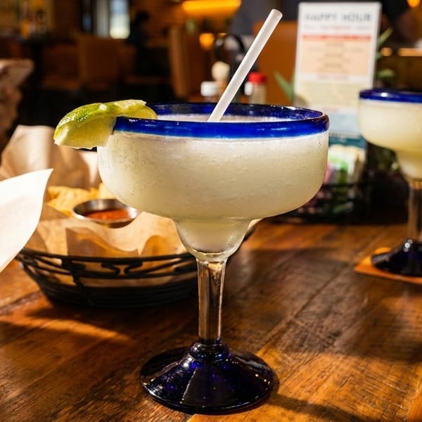 Two frozen margaritas with lime wedges and a basket of tortilla chips on a wooden table.