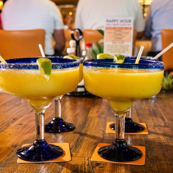 Two frozen margaritas with lime wedges and straws on a wooden table in a restaurant.