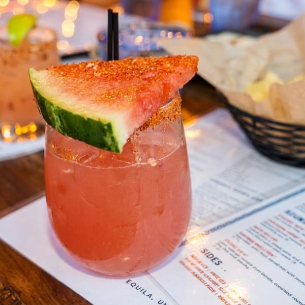 Watermelon margarita with a chili-lime rim and a basket of chips on a wooden table.