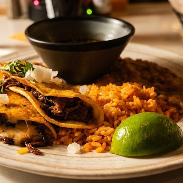 Birria tacos with rice, beans, a lime wedge, and a bowl of consommé on a plate.