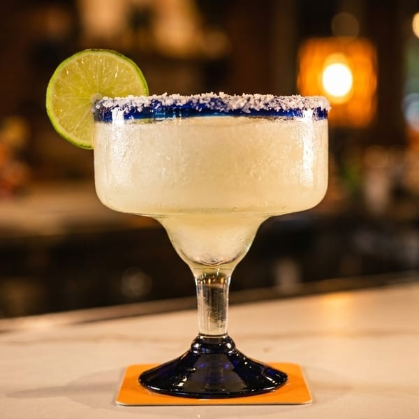 A frozen margarita with a salted rim and lime wedge sits on a bar.
