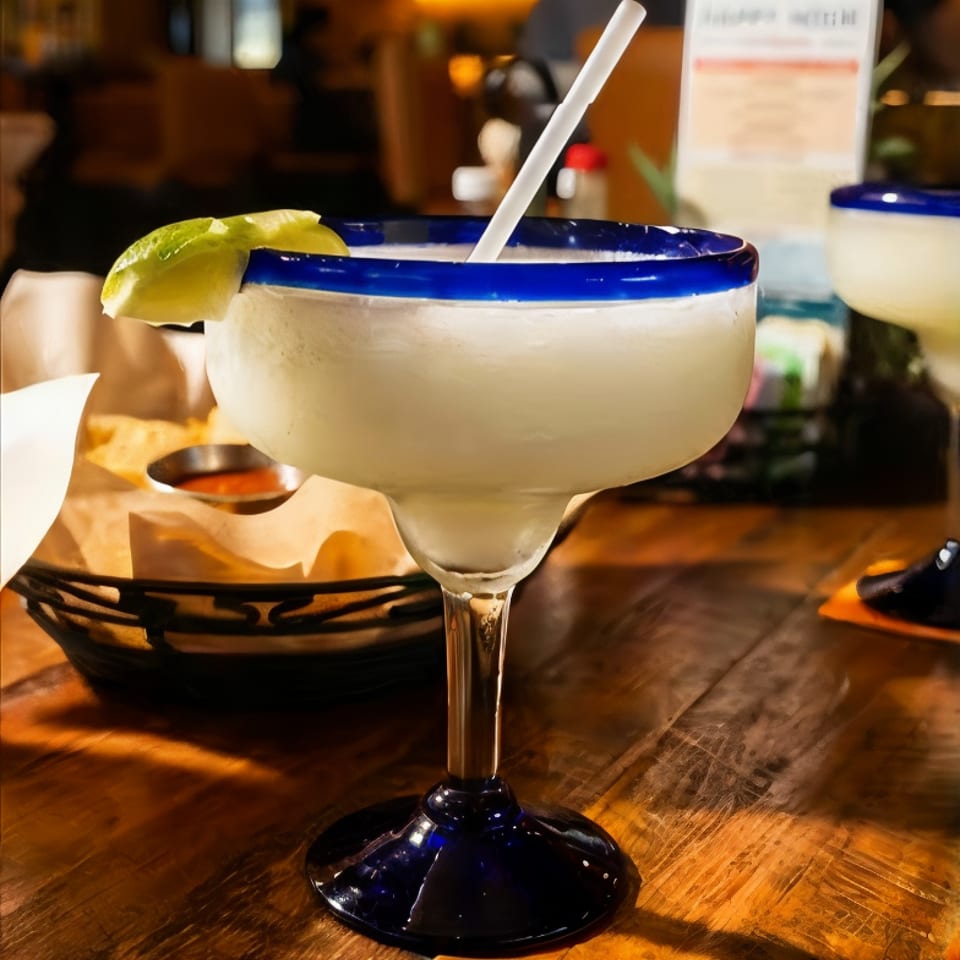 Frozen margarita in a blue-rimmed glass garnished with lime, served on a wooden restaurant table.